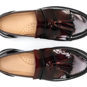 May include: Pair of glossy, burgundy leather loafers with decorative tassels. The shoes have a classic design with a rounded toe and a stitched sole. The interior is a light tan color.