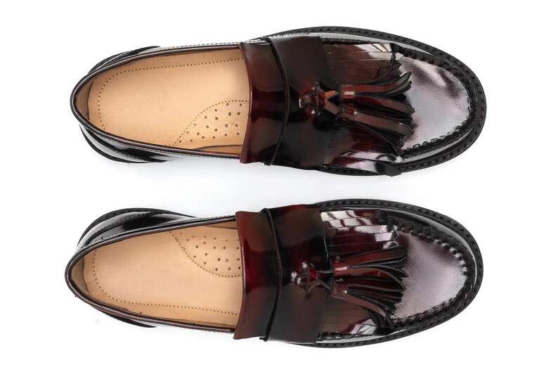 May include: Pair of glossy, burgundy leather loafers with decorative tassels. The shoes have a classic design with a rounded toe and a stitched sole. The interior is a light tan color.