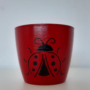 Puede incluir: Una pequeña maceta de madera roja con un diseño de mariquita negra. La mariquita está centrada en la parte delantera de la maceta y tiene cabeza, cuerpo y puntos negros. La maceta es cilíndrica y tiene un acabado liso.