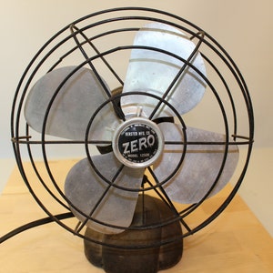 Puede incluir: Un ventilador eléctrico antiguo con una jaula y aspas de metal negro. El ventilador tiene una base oscura y una etiqueta que dice "ZERO" y "MODEL 12500". Está sobre una superficie de madera clara.