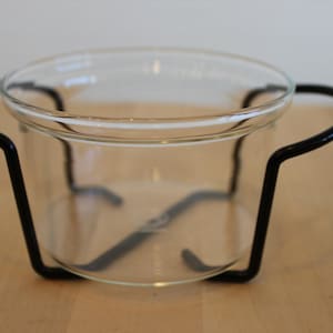 Puede incluir: Un cuenco de vidrio transparente con un soporte de metal negro. El cuenco es redondo y tiene una superficie lisa. El soporte está hecho de metal delgado y tiene un diseño simple.