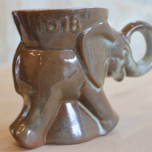 May include: A brown ceramic mug shaped like an elephant with the year 1978 embossed on the side. The mug has a glazed finish and a handle shaped like an elephant's trunk.
