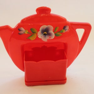 May include: Red plastic teapot-shaped kitchen utensil holder with a floral design. The holder has a small compartment for storing utensils.