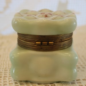May include: A small, pale green ceramic trinket box with a hinged lid. The lid has a raised, scalloped design with a subtle floral pattern in shades of pink and white. The box has a brass hinge and clasp.