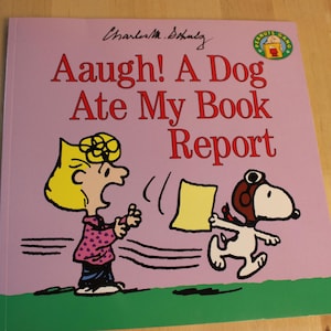 Pode incluir: Um livro infantil quadrado com capa roxa clara apresentando os personagens Peanuts, Sally Brown e Snoopy. O título Aaugh! A Dog Ate My Book Report está em vermelho. Um logotipo circular Peanuts Gang está no canto superior direito.