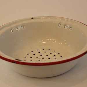 Puede incluir: Un colador de esmalte blanco con borde rojo. El colador tiene una serie de pequeños agujeros en la base y varios agujeros más grandes alrededor del borde. Este utensilio de cocina es ideal para escurrir pasta o lavar verduras.