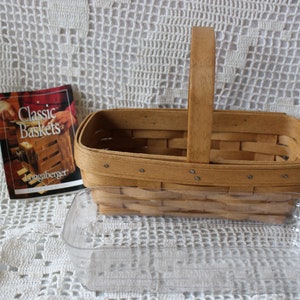 Peut inclure: Un panier tissé brun clair avec une poignée en bois, un plat en verre rectangulaire transparent et une brochure intitulée "Classic Baskets". Le panier est rectangulaire avec un design tissé et des rivets métalliques. La brochure a une couverture rouge et marron.