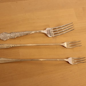 May include: Three silver-toned metal forks arranged horizontally. The top fork is wide with an ornate floral handle. The middle fork is slender with a decorative handle. The bottom fork is thin and plain with a smooth, tapered handle design.