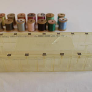 May include: A clear, rectangular sewing box with a yellow tint, containing a variety of colorful thread spools. The box has a hinged lid and metal closures. The spools are in various colors, including green, blue, pink, and brown.