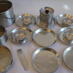 Puede incluir: Una colección de utensilios de cocina de camping de aluminio vintage, que incluye ollas, tazas, platos y un recipiente con tapa. Los artículos están dispuestos sobre una superficie blanca, mostrando su diseño simple y utilitario.