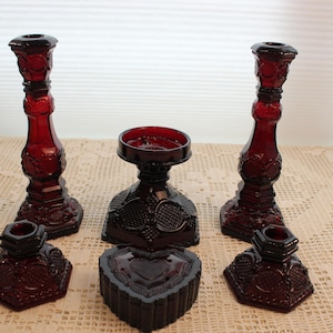 May include: A collection of six deep red pressed glass items including two tall candlesticks, two short votive holders, one central pedestal bowl, and one heart-shaped lidded dish, all featuring intricate geometric and floral patterns.
