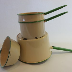 May include: A stack of vintage beige enamelware pots with green handles and trim. The set includes three pots of varying sizes, one lying on its side. The pots have a classic, retro design.