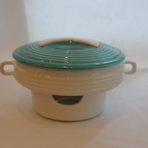 May include: A vintage ceramic casserole dish. It has a teal lid and a cream-coloured base. The lid features concentric ridges and a cream-coloured handle. The dish has two small handles and a circular opening on the base. A classic serving piece.