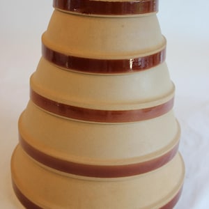 Op de afbeelding: Een stapel beige keramische mengkommen met een donkerbruine band rond elke kom. De kommen zijn genest, met de grootste kom onderaan en de kleinste bovenaan. De kommen hebben een gladde, glanzende afwerking. Geschikt voor de keuken.