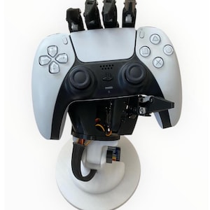 Könnte beinhalten: Ein weißer und schwarzer Roboterarm mit einem PlayStation 5-Controller. Der Controller hat das Standard-Tastenlayout und ist auf einer kreisförmigen Basis montiert. Der Roboterarm hat schwarze Finger.