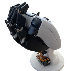Könnte beinhalten: Eine Roboterhand mit schwarzen und weißen Komponenten, einschließlich beweglicher Finger und einer weißen Basis. Die Hand ist für das Halten eines Controllers konzipiert, mit sichtbarer Verkabelung und dem Text "POZER (SV2)" auf der Basis.