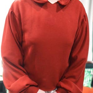 May include: A red sweatshirt with a unique collar and long sleeves. The collar has a rounded shape. The sleeves are gathered at the cuffs. The fabric appears to be a soft material.