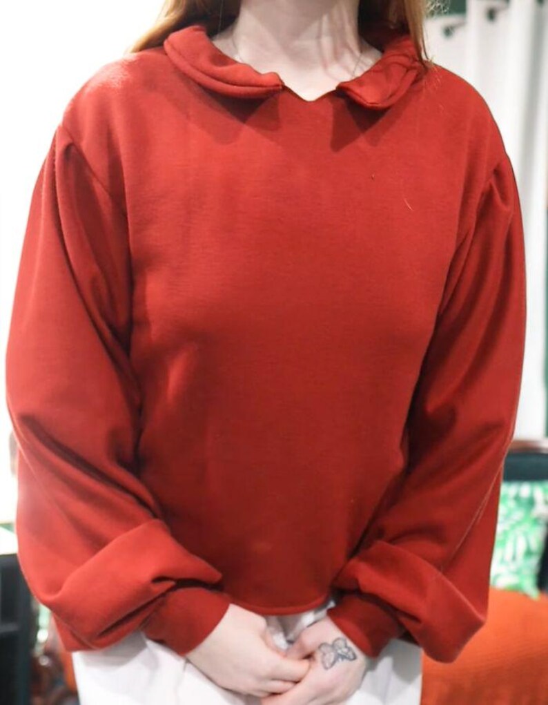 May include: A red sweatshirt with a unique collar and long sleeves. The collar has a rounded shape. The sleeves are gathered at the cuffs. The fabric appears to be a soft material.