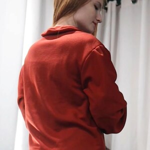 May include: A red sweater with a high collar and long sleeves. The sweater is made of a soft material and is paired with white pants. The model is facing away from the camera, with their head turned to the side.