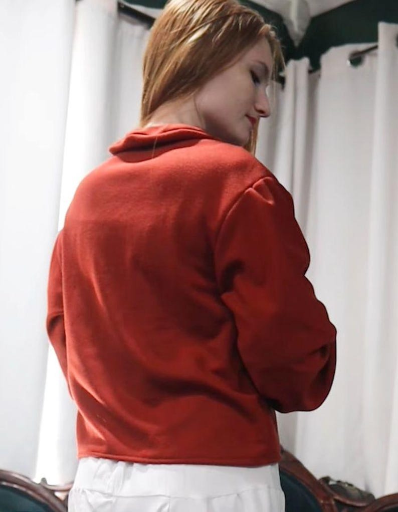 May include: A red sweater with a high collar and long sleeves. The sweater is made of a soft material and is paired with white pants. The model is facing away from the camera, with their head turned to the side.