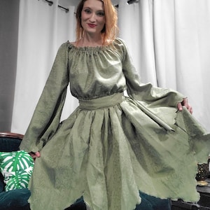 Puede incluir: Un vestido verde salvia con mangas largas y una falda acampanada. El vestido presenta un escote fruncido y una cintura entallada. La tela tiene un sutil estampado floral. La falda tiene un dobladillo asimétrico único. El vestido es de un material suave y fluido.