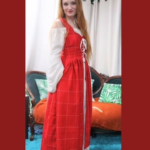 Peut inclure: Une robe rouge à carreaux avec un chemisier blanc en dessous. La robe a un col carré et un détail de laçage sur le devant. Les manches du chemisier sont longues et bouffantes. La robe arrive au genou et a une légère forme trapèze.