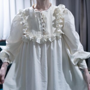 May include: An off-white long-sleeve dress featuring a button-up front placket with vertical pleats, a round neckline, and ruffled trim detailing along the chest and shoulders. The garment has loose, voluminous sleeves with cuffs.