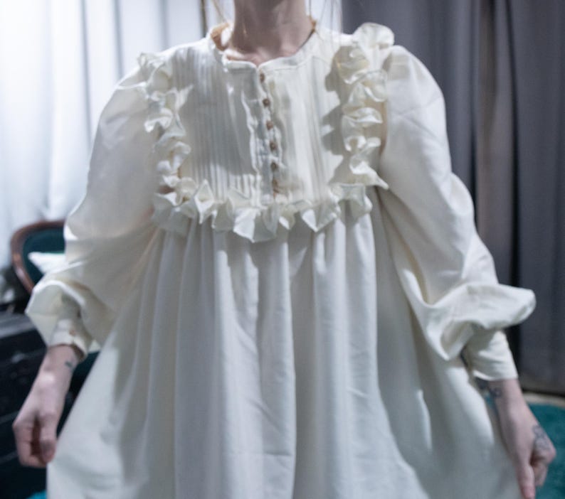 May include: An off-white long-sleeve dress featuring a button-up front placket with vertical pleats, a round neckline, and ruffled trim detailing along the chest and shoulders. The garment has loose, voluminous sleeves with cuffs.