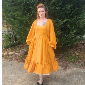 Könnte beinhalten: Senfgelbes Kleid mit langen, voluminösen Ärmeln und einem quadratischen Ausschnitt. Das Kleid hat einen gerüschten Saum und wird mit schwarzen Mary Jane-Schuhen getragen. Das Kleid scheint aus einem weichen Stoff zu sein.