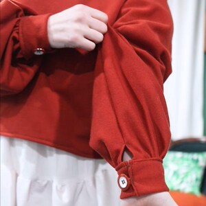 May include: A red long-sleeved top with a round neckline and buttoned cuffs. The sleeves are gathered at the shoulders and cuffs. The top is paired with a white skirt. The buttons are white with a dark number 11.