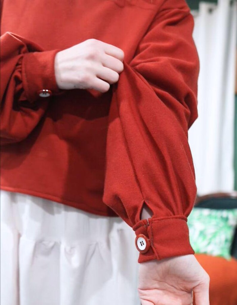 May include: A red long-sleeved top with a round neckline and buttoned cuffs. The sleeves are gathered at the shoulders and cuffs. The top is paired with a white skirt. The buttons are white with a dark number 11.