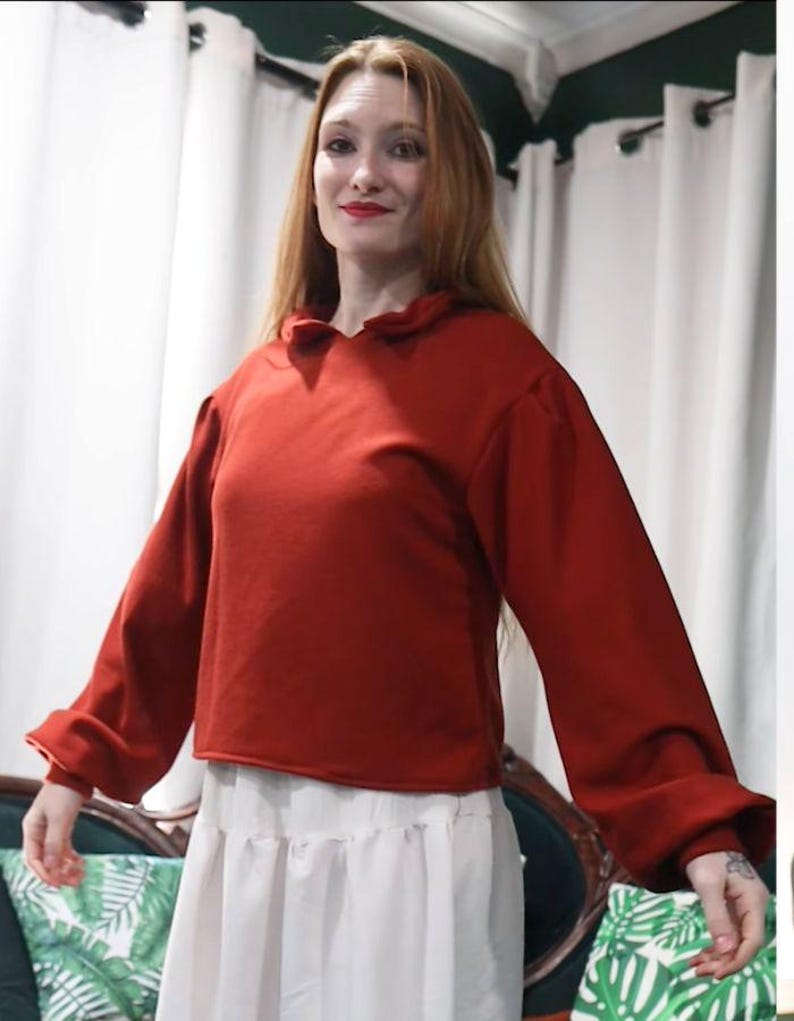 May include: A rust-colored long-sleeved top with a ruffled collar and a cropped hem. The sleeves are gathered at the cuffs. The top is paired with a white skirt.
