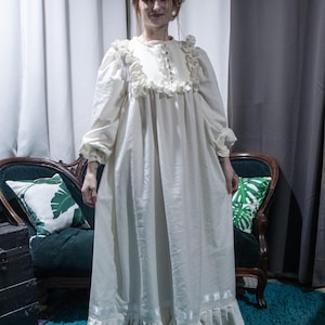 May include: A floor-length cream-colored dress featuring long sleeves with gathered cuffs, a high neckline with a button-down placket, and ruffled detailing across the chest and hem. The garment includes a horizontal ribbon trim near the bottom.