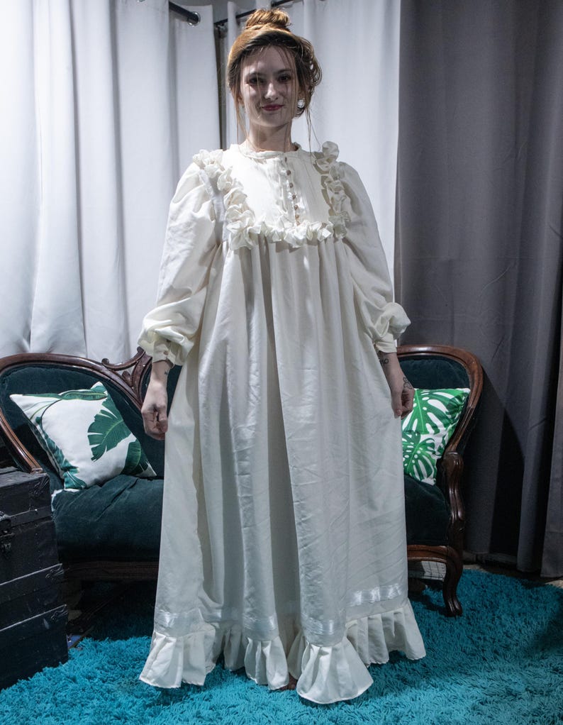 May include: A floor-length cream-colored dress featuring long sleeves with gathered cuffs, a high neckline with a button-down placket, and ruffled detailing across the chest and hem. The garment includes a horizontal ribbon trim near the bottom.