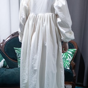 May include: A floor-length, off-white nightgown featuring long, full sleeves with gathered cuffs, a high neckline, and a pleated bodice. The hem is finished with a wide, ruffled trim that includes a horizontal ribbon detail near the bottom edge.