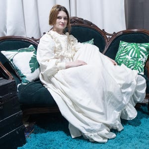 May include: A floor-length cream-colored dress with long sleeves, a ruffled bodice, and a high neckline. The garment features a full, flowing skirt with a ruffled hemline, constructed from a solid, opaque fabric with a matte finish.