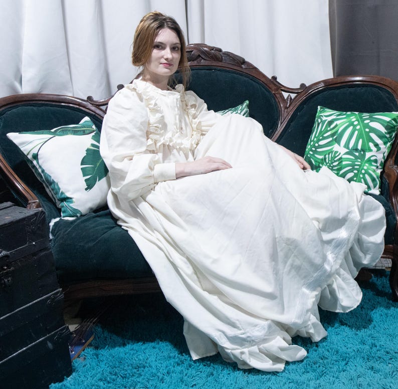 May include: A floor-length cream-colored dress with long sleeves, a ruffled bodice, and a high neckline. The garment features a full, flowing skirt with a ruffled hemline, constructed from a solid, opaque fabric with a matte finish.