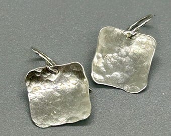 May include: A pair of silver-toned earrings. Each earring features a hammered, square-shaped design with a textured surface. The earrings have a simple hook closure. The background is a solid gray.