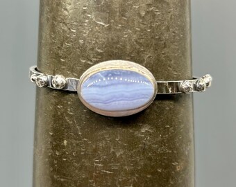 May include: A silver bracelet featuring an oval, light blue gemstone. The gemstone is set in a silver bezel. The bracelet has a simple band with small, round silver accents on each side of the gemstone.