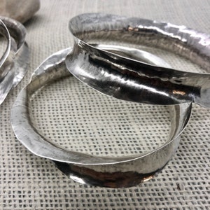 May include: Three silver bracelets with a hammered finish. The bracelets are stacked on top of each other and are lying on a textured fabric surface.