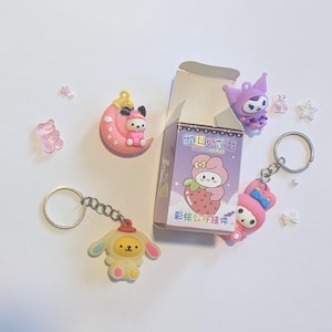 May include: A collection of colorful keychains and small figurines. The keychains feature cartoon characters in pink, yellow, and blue. A box with Japanese text and a strawberry character is also present, along with decorative accents.