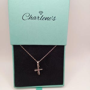 Peut inclure: Un collier avec un pendentif en forme de croix argentée, orné d'un petit cœur rouge, présenté dans un coffret bleu clair ouvert. L'intérieur du coffret est noir. Le couvercle porte l'inscription "Charlene's".