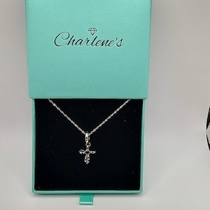 Peut inclure: Un collier avec pendentif en forme de croix argentée présenté dans un coffret cadeau turquoise ouvert. Le coffret révèle le collier sur un fond noir. Le couvercle du coffret porte l'inscription "Charlene's".