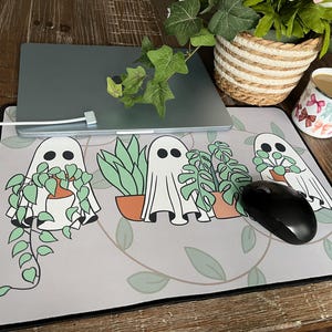 May include: A grey mouse mat with a quirky design of cartoon ghosts holding potted plants. A black computer mouse sits on the right. A laptop rests on top of the mat. The mat has a black border.