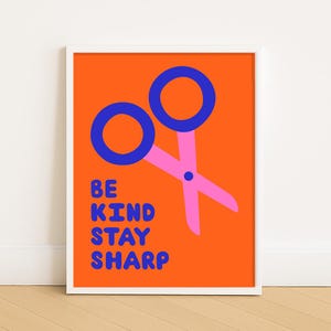以下が含まれることがあります： オレンジ色の背景に、ピンクのはさみと青い持ち手が描かれたフレーム入りアートプリント。「BE KIND STAY SHARP」の文字が青色で書かれています。モダンでミニマリストなデザインの作品です。