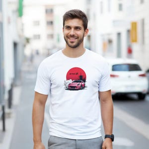 May include: White t-shirt featuring a graphic of a pink sports car with the text "370Z" in front of a red sun and Japanese-style trees. The shirt is a crew neck and short-sleeved.
