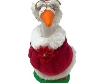 May include: A plush goose figurine wearing a red sweater with a white faux fur collar and a red fabric poinsettia accent. The goose has an orange beak, wears gold-rimmed round glasses, and has a small red hat on its head. It stands on a green base.