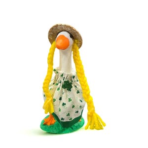 May include: A novelty goose ornament, wearing a straw hat and long yellow plaits. The goose is dressed in a white dress with green shamrocks and stands on a green base. It has an orange beak and feet.