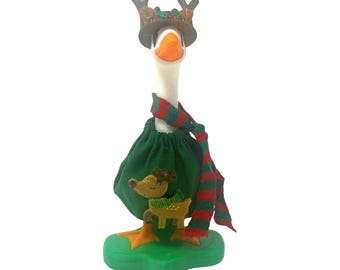 May include: A white goose figurine wearing a brown hat with reindeer antlers, a green fabric dress with a small reindeer appliqué, and a red and green striped knit scarf. The goose stands on a green base and has an orange beak and orange feet.