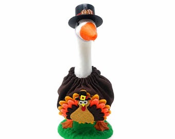 May include: A white goose figurine wearing a black top hat with a small pumpkin accent and a brown fabric cape. The cape features a felt turkey applique with colorful orange and red feathers, a yellow beak, and a tiny black hat on its head.